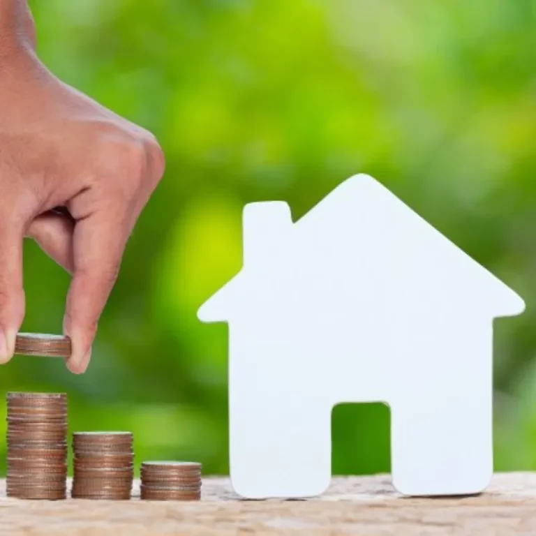 Biały domek na zielonym tle z monetami układanymi przez jakąś osobę symbolizujący oszczędności w ogrzewaniu z pompami ciepła Daikin